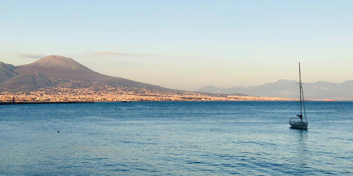 Vue panoramique sur la mer Méditerranée avec le mont Vésuve et la ville de Naples au coucher du soleil, un voilier seul sur l'eau.