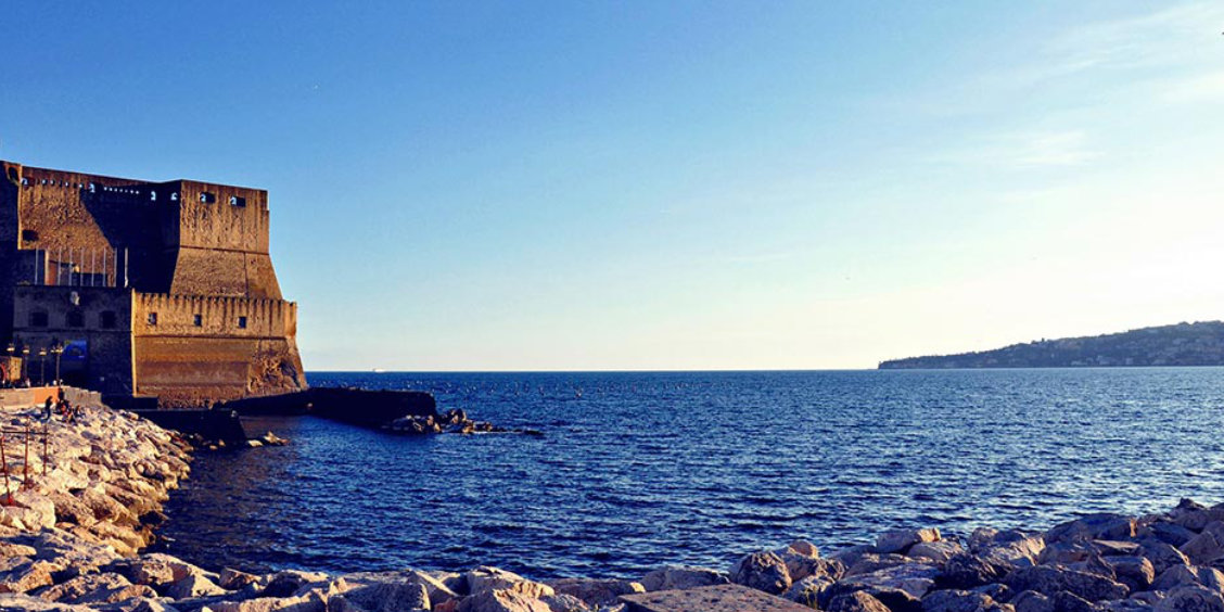 Le Castel dell'Ovo en bord de mer à Naples avec les rochers du littoral au premier plan.