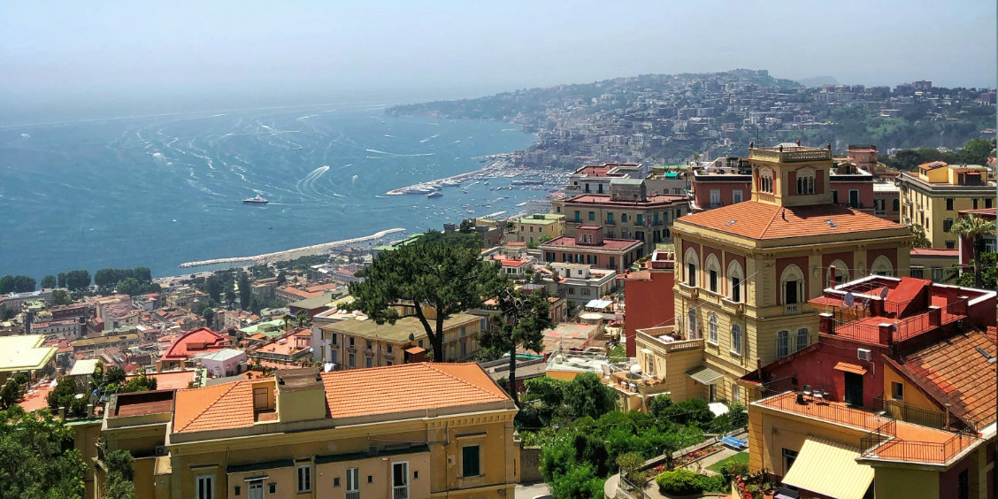Vue panoramique sur les toits colorés de la ville de Naples et sa vaste baie parsemée de bateaux sous un ciel lumineux.