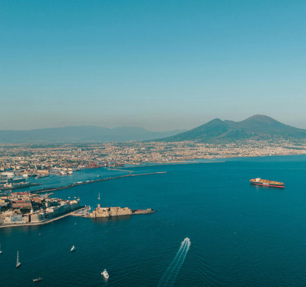Panorama aérien sur le golfe de Naples avec le Vésuve en arrière-plan et le Castel dell'Ovo sur l'eau bleue.