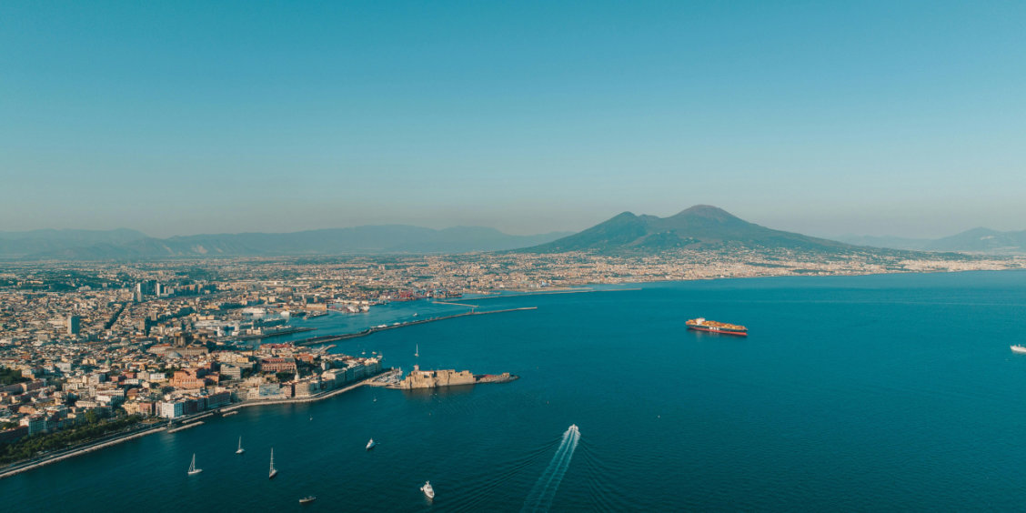 Panorama aérien sur le golfe de Naples avec le Vésuve en arrière-plan et le Castel dell'Ovo sur l'eau bleue.