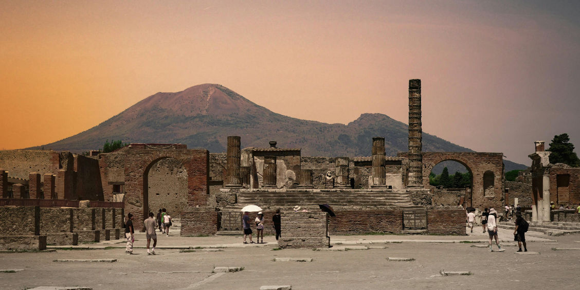 Vue panoramique des ruines du forum de Pompéi avec le mont Vésuve en arrière-plan sous un ciel orangé.
