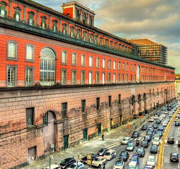 Long bâtiment historique aux façades rouges à Naples, bordant une avenue animée avec de nombreuses voitures et des arbres sur le côté.