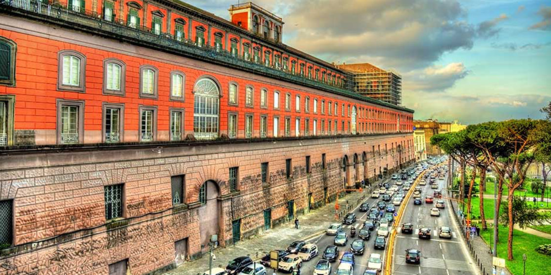 Long bâtiment historique aux façades rouges à Naples, bordant une avenue animée avec de nombreuses voitures et des arbres sur le côté.