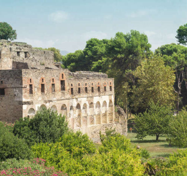 Bâtiment antique en pierre et brique avec des arcades dans les ruines de Pompéi, entouré d'une végétation verdoyante et de fleurs roses.