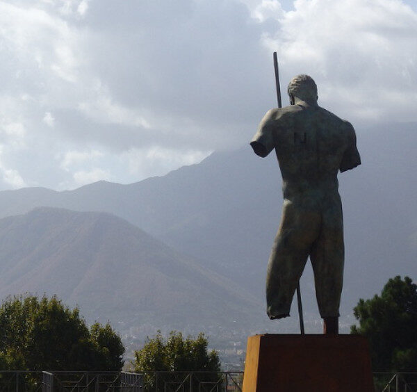 Statue de bronze à Pompéi sous un ciel gris avec vue sur les montagnes et les ruines antiques.