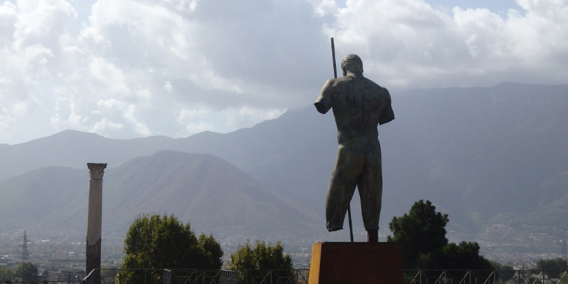 Statue de bronze à Pompéi sous un ciel gris avec vue sur les montagnes et les ruines antiques.