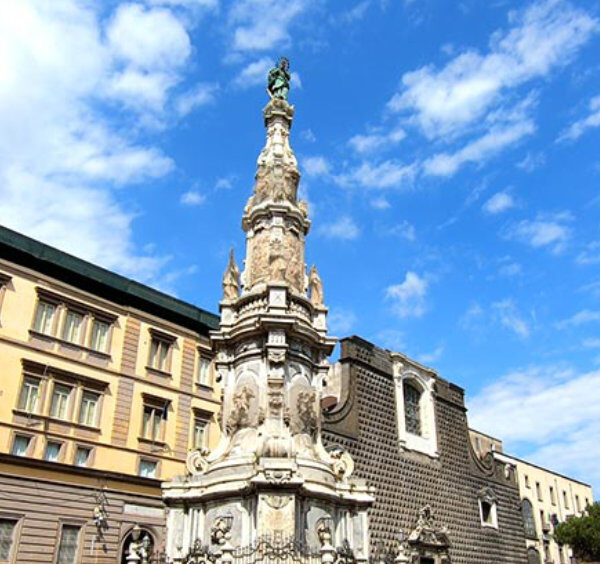 Piazza del Gesù Nuovo à Naples avec l’obélisque baroque et l’église en arrière-plan sous un ciel bleu.