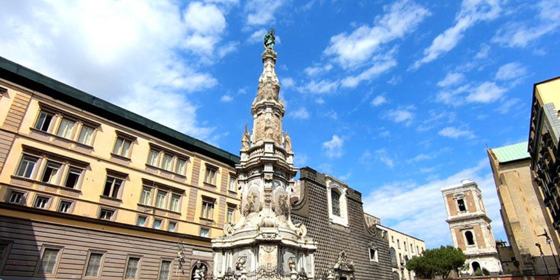 Piazza del Gesù Nuovo à Naples avec l’obélisque baroque et l’église en arrière-plan sous un ciel bleu.