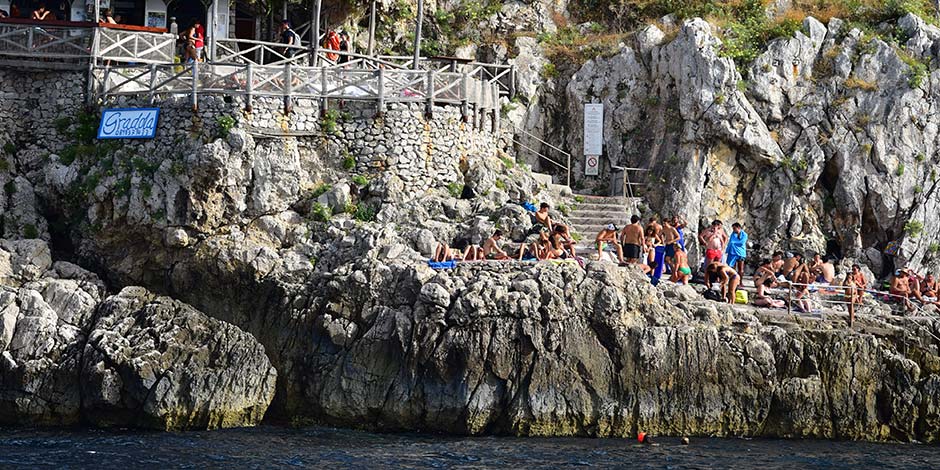 Baigneurs installés sur les rochers de la plage de Gradola à Capri, au pied d'une terrasse en bois.