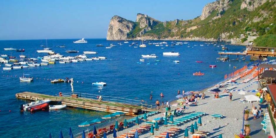 Plage de Marina del Cantone à Nerano avec bateaux, parasols et falaises en arrière-plan.