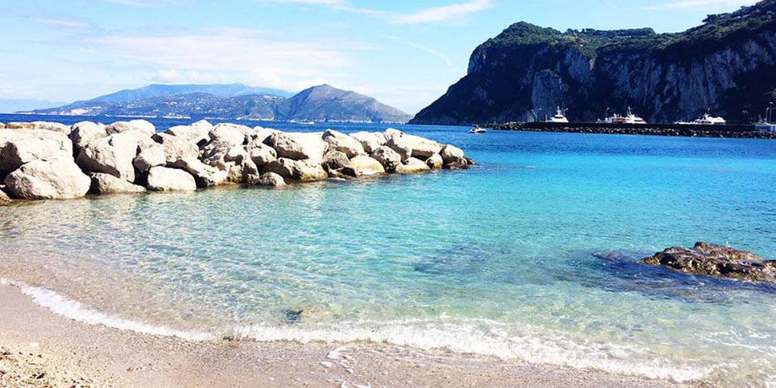 Eaux turquoise et plage de galets à Marina Grande, Capri, entourée de rochers blancs sous un ciel dégagé.