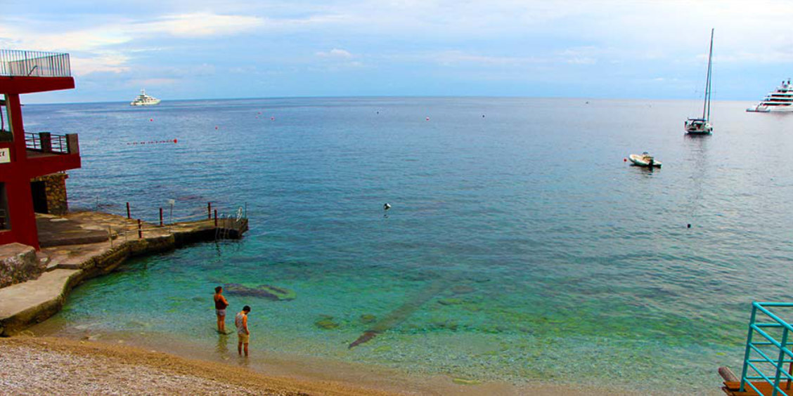 Plage de galets à Marina Piccola à Capri avec des eaux cristallines, deux personnes au bord de l'eau et des bateaux à l'horizon.