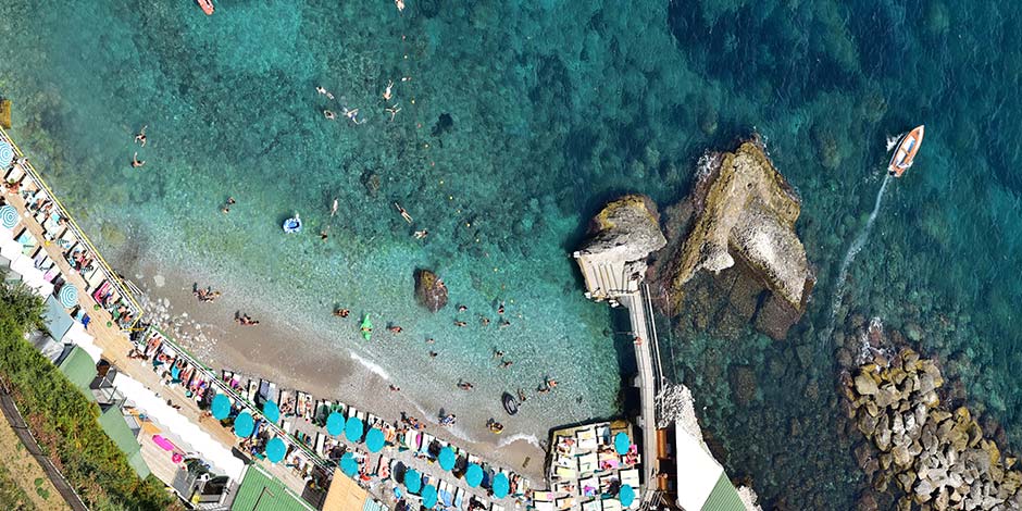 Vue aérienne de la plage de Palazzo a Mare à Capri avec ses eaux turquoise, ses parasols bleus et son ponton caractéristique.