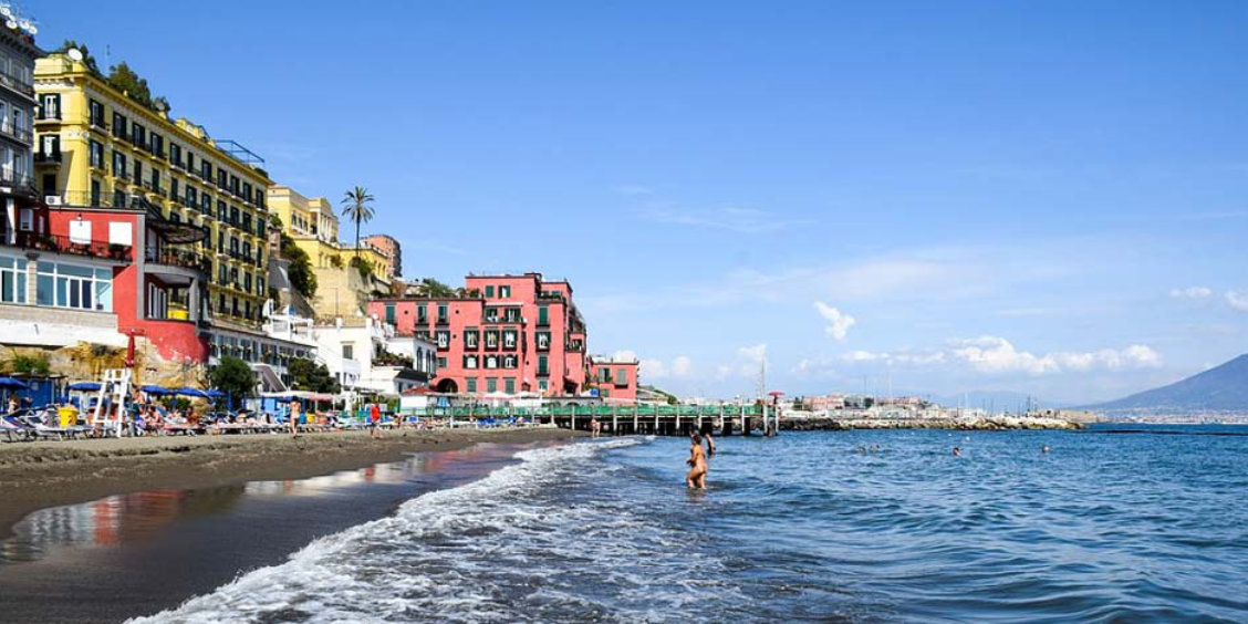 Plage de Naples avec maisons colorées, mer calme et vue sur le Vésuve le long du littoral.