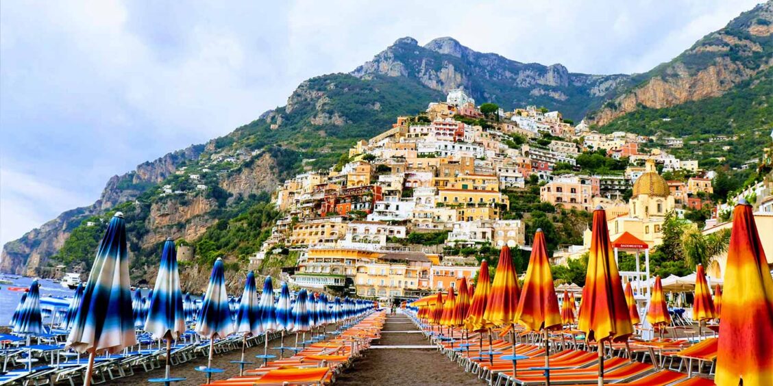 Plage de Positano avec rangées de parasols colorés et maisons accrochées à la montagne.