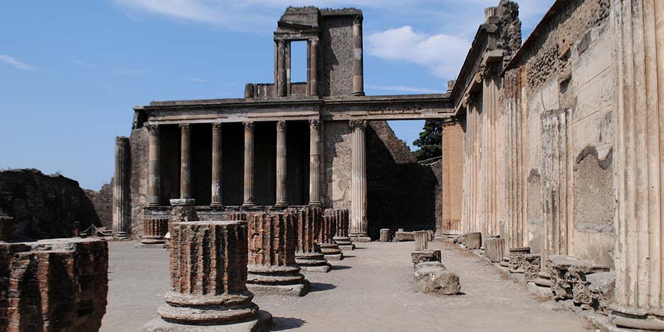 Ruines antiques de Pompéi avec des colonnes imposantes et des vestiges de structures en pierre sous un ciel bleu.