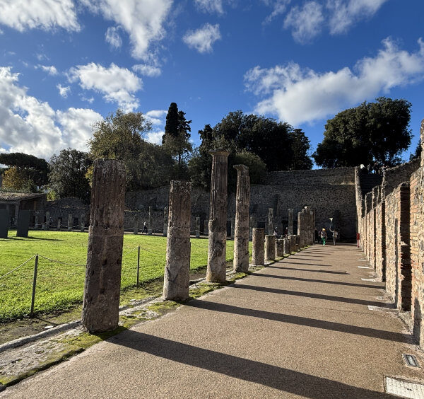 Allée dans les ruines de Pompéi bordée de colonnes antiques en pierre et de murs en briques, sous un ciel bleu nuageux.