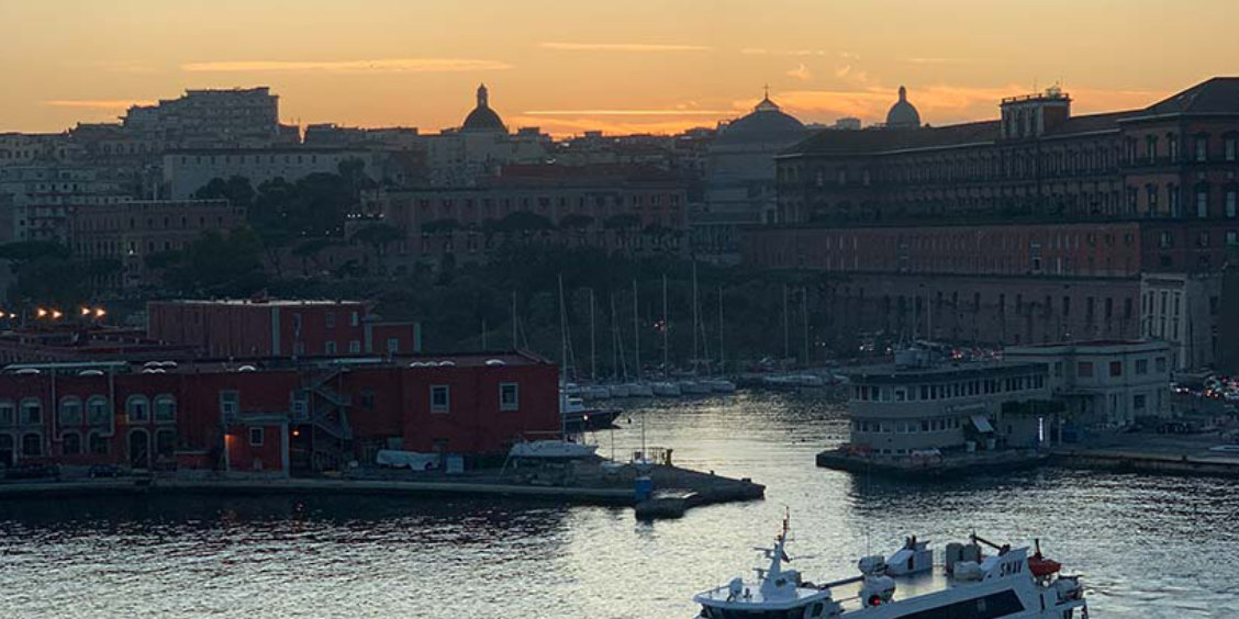 Vue panoramique du port de Naples au coucher du soleil avec le Palais Royal et un ferry SNAV au départ du Molo Beverello.