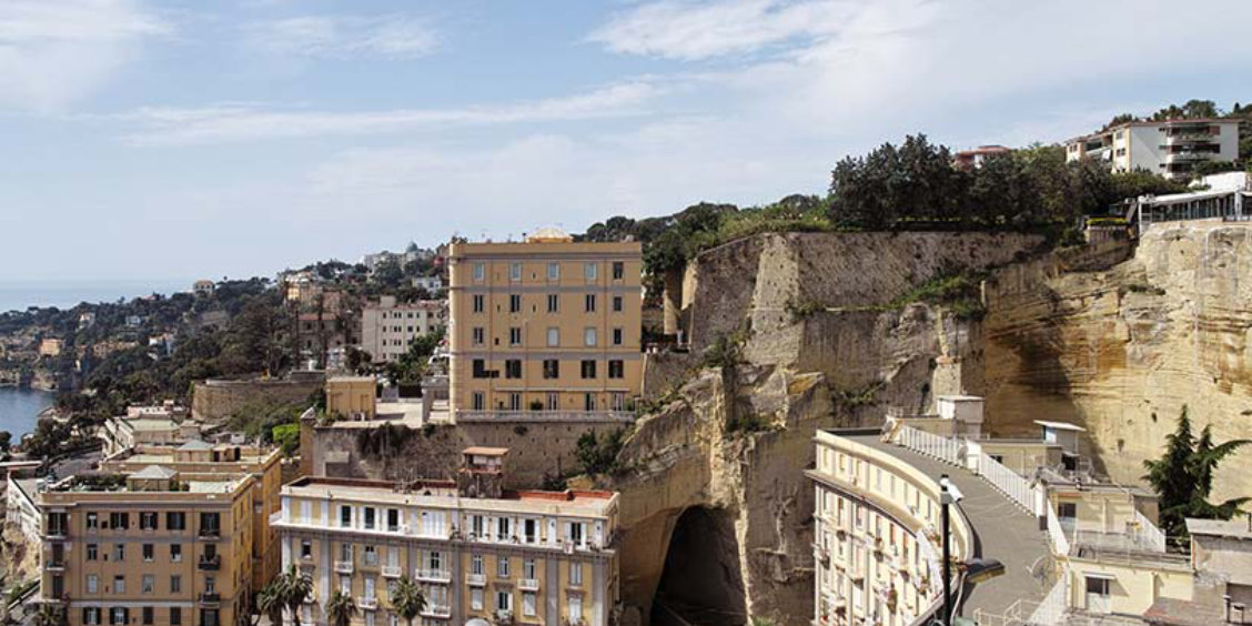 Vue aérienne de Posillipo à Naples, montrant des bâtiments jaunes accrochés aux imposantes falaises de tuf avec la mer Tyrrhénienne en arrière-plan et l'entrée d'un tunnel.