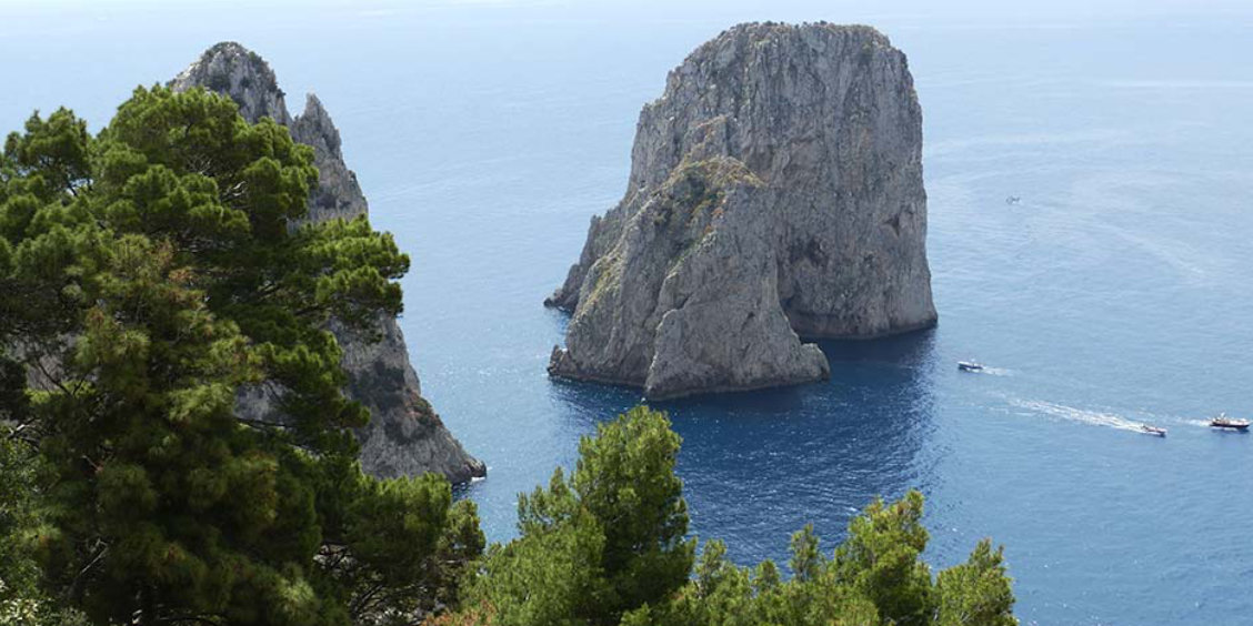 Vue des rochers Faraglioni à Capri à travers les pins avec des bateaux naviguant sur la mer bleue.