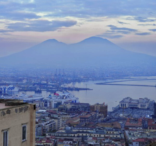 Vue panoramique de Naples avec le Vésuve en arrière-plan et le port sous un ciel nuageux.