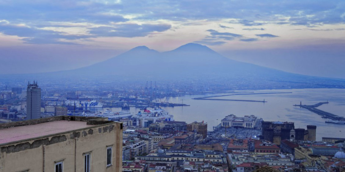 Vue panoramique de Naples avec le Vésuve en arrière-plan et le port sous un ciel nuageux.