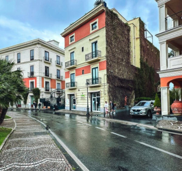 Rue de Sorrente sous la pluie avec façades colorées, palmiers et chaussée mouillée.