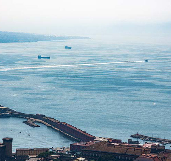 Vue aérienne du port de Naples avec le Castel Nuovo et la silhouette du Vésuve à l'horizon.