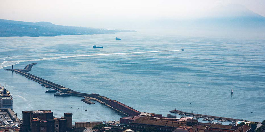 Vue aérienne du port de Naples avec le Castel Nuovo et la silhouette du Vésuve à l'horizon.