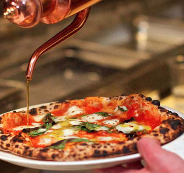 Un filet d'huile d'olive versé avec précision depuis une burette traditionnelle en cuivre sur une pizza Margherita fraîchement sortie du four.
