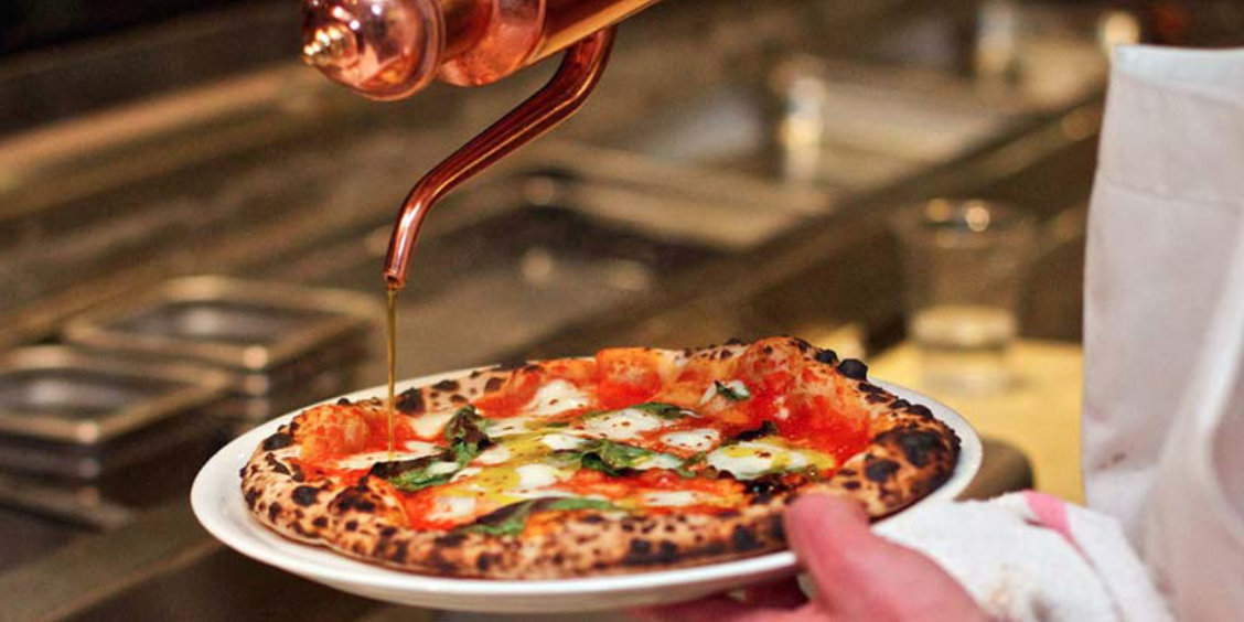 Un filet d'huile d'olive versé avec précision depuis une burette traditionnelle en cuivre sur une pizza Margherita fraîchement sortie du four.
