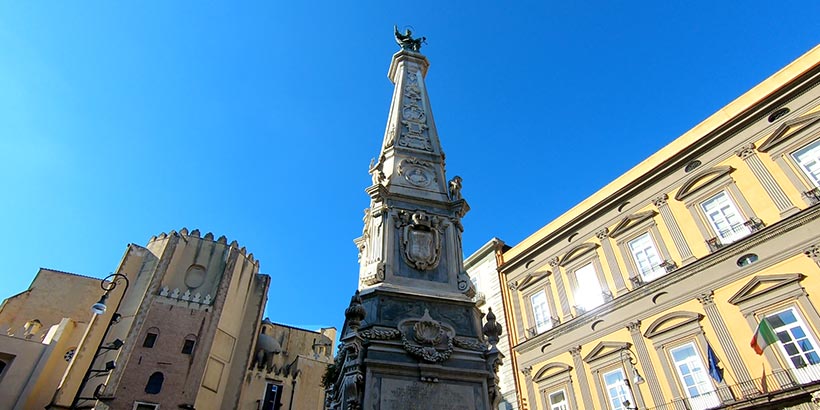 Obélisque de San Domenico Maggiore à Naples avec église et bâtiments historiques sous un ciel bleu.