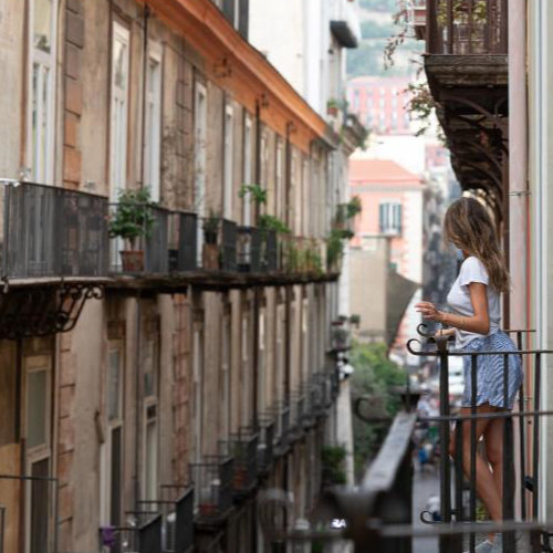 Jeune femme sur un balcon typique de Naples, admirant une ruelle étroite bordée de bâtiments historiques.