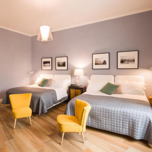 Chambre d'hôtel moderne et spacieuse avec deux lits doubles, des murs gris-mauve et deux fauteuils jaune vif.