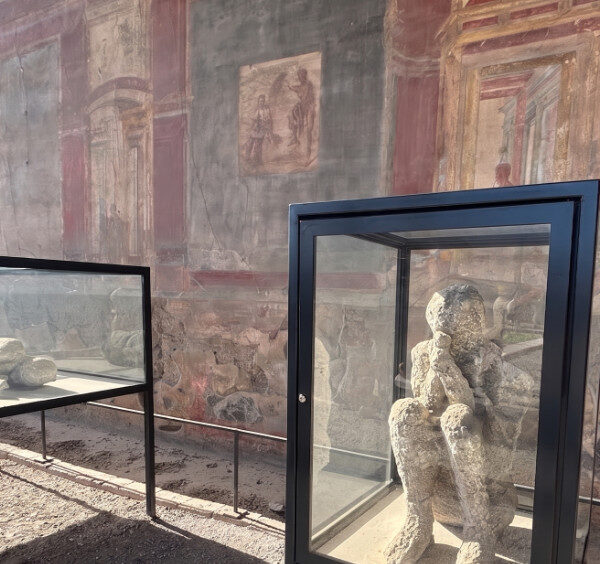Exposition en plein air à Pompéi : moulages en plâtre des victimes de l'éruption de 79 apr. J.-C. dans des vitrines, devant un mur à fresques romain.
