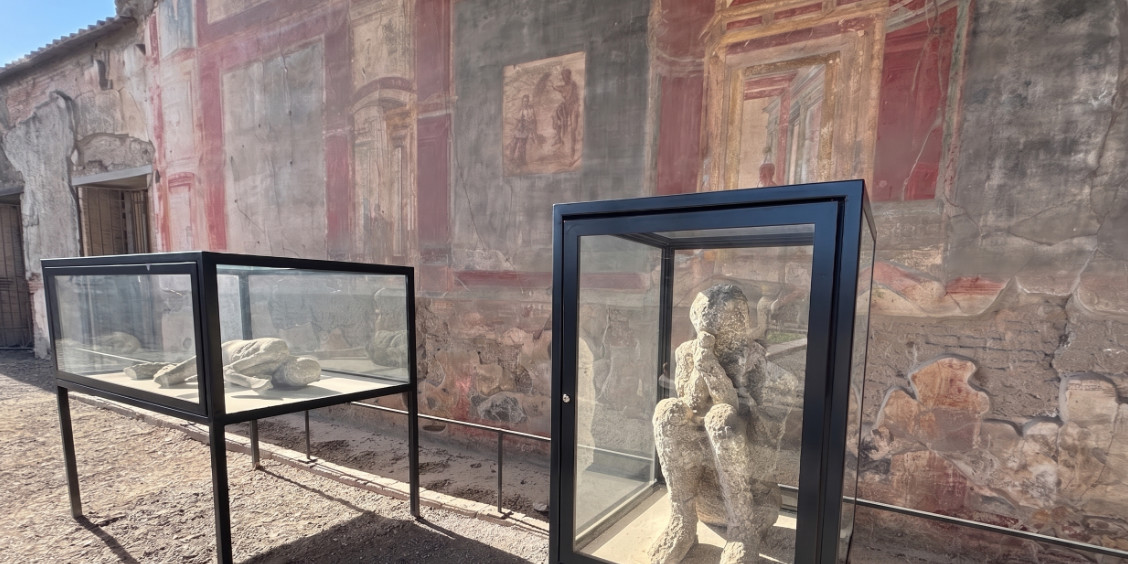 Exposition en plein air à Pompéi : moulages en plâtre des victimes de l'éruption de 79 apr. J.-C. dans des vitrines, devant un mur à fresques romain.