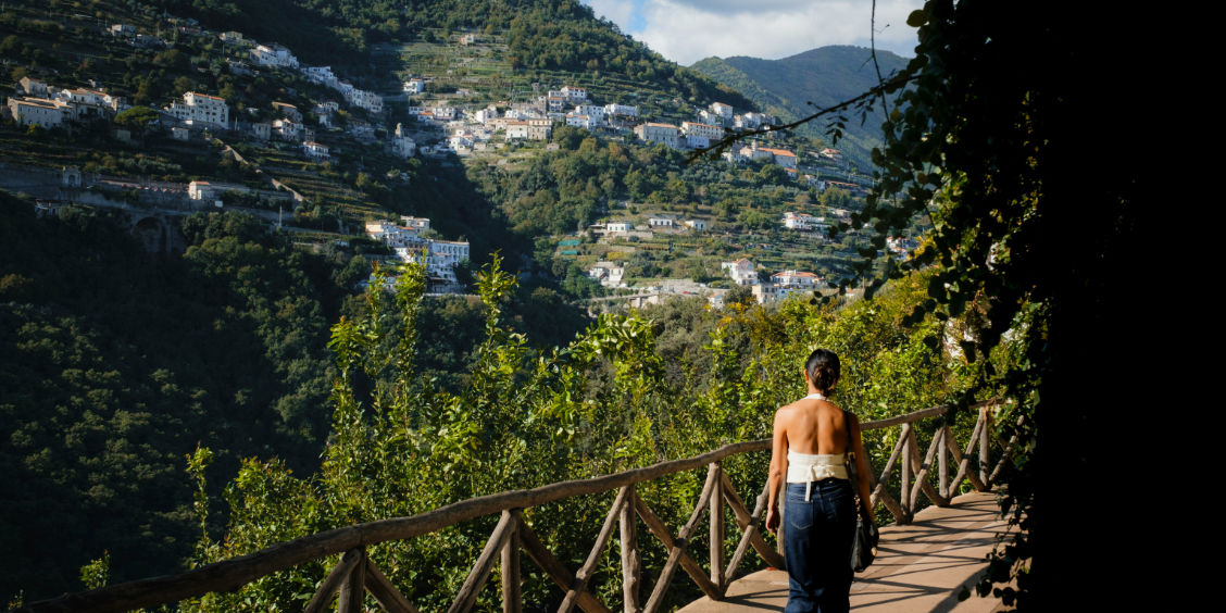 Promeneuse sur un sentier pavé bordé d'une barrière en bois face aux collines verdoyantes et villages perchés de Ravello.
