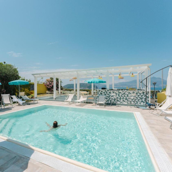 Piscine extérieure ensoleillée avec vue sur la mer, transats et bar ombragé sous une pergola.