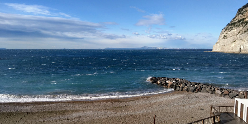 Plage de Sorrente en février avec mer agitée, jetée rocheuse et falaise côtière.