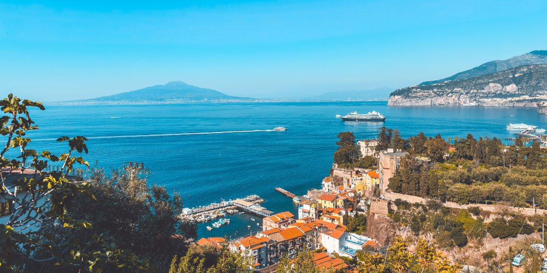 Vue panoramique de la ville de Sorrente bâtie sur les falaises, surplombant un petit port et la mer avec la silhouette du mont Vésuve à l'horizon.