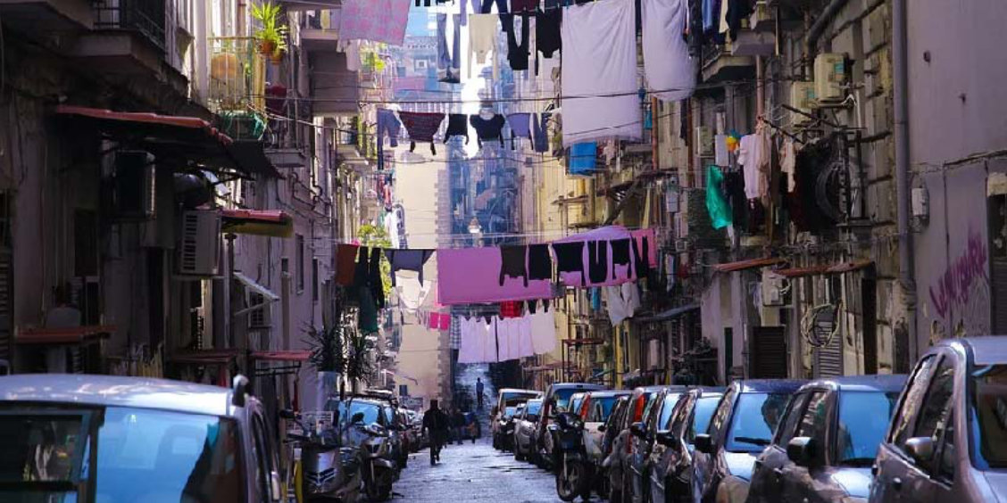 Rue étroite typique de Naples avec voitures garées des deux côtés, linge suspendu entre les immeubles et ambiance de quartier populaire.