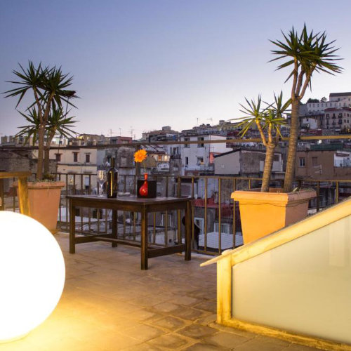 Terrasse panoramique sur le toit de Naples au crépuscule, aménagée avec des plantes vertes et une table pour se détendre face à la ville.