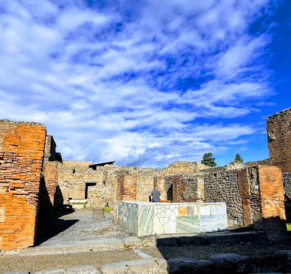 Vue des ruines d'un Thermopolium à Pompéi montrant des comptoirs en marbre et des murs en briques rouges sous un ciel bleu parsemé de nuages blancs.