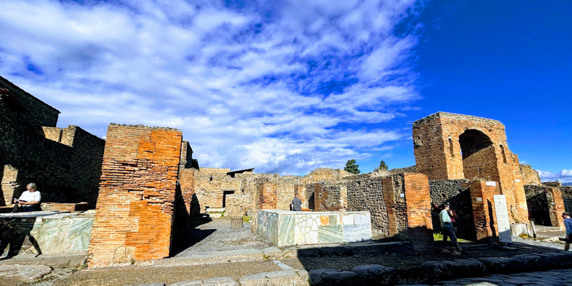 Vue des ruines d'un Thermopolium à Pompéi montrant des comptoirs en marbre et des murs en briques rouges sous un ciel bleu parsemé de nuages blancs.