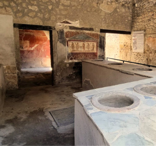 Comptoir en pierre d'un ancien thermopolium romain avec des ouvertures circulaires pour les jarres de nourriture et une fresque murale en arrière-plan.