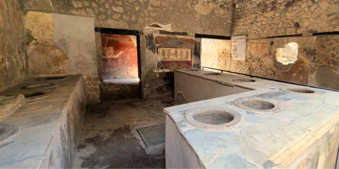 Comptoir en pierre d'un ancien thermopolium romain avec des ouvertures circulaires pour les jarres de nourriture et une fresque murale en arrière-plan.