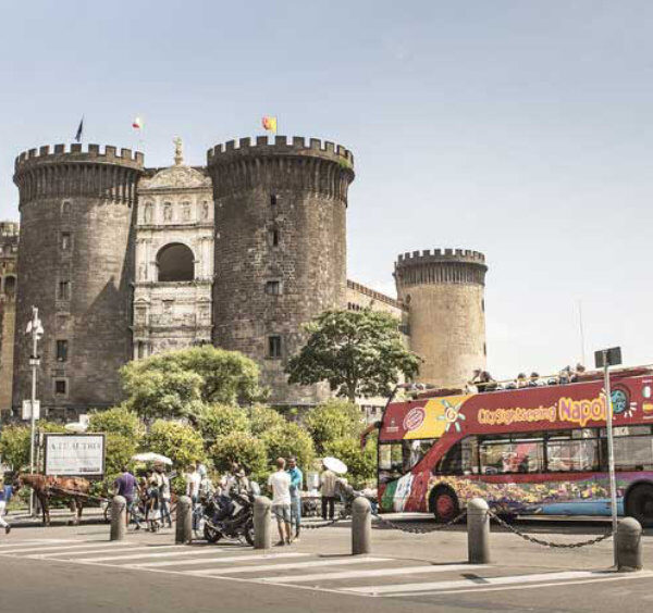 Bus touristique à Naples près du Castel Nuovo, avec visiteurs et circulation en centre-ville.