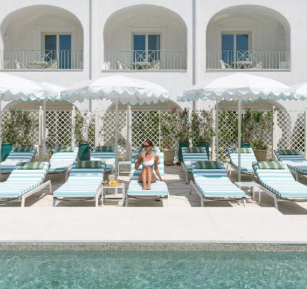 Piscine d'un hôtel à Capri avec transats rayés bleu et blanc, parasols et façade méditerranéenne immaculée.