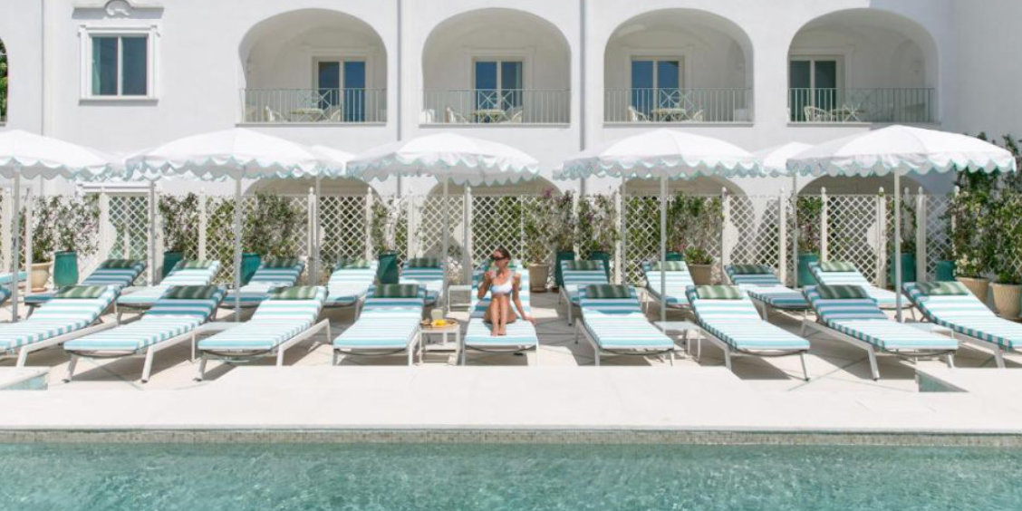 Piscine d'un hôtel à Capri avec transats rayés bleu et blanc, parasols et façade méditerranéenne immaculée.